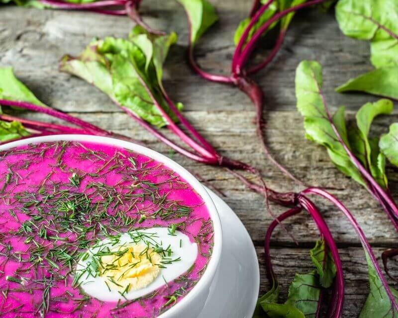 Na stole leży miska pełna różowego, schłodzonego chłodnika z buraczków, z jajkiem i szczypiorkiem, a obok leżą liście buraka - botwina.