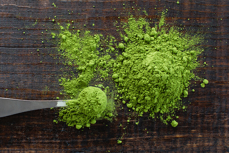 Na drewnianej ławie rozsypano zieloną chlorellę, która przebiera z metalowej łyżki - miarki.