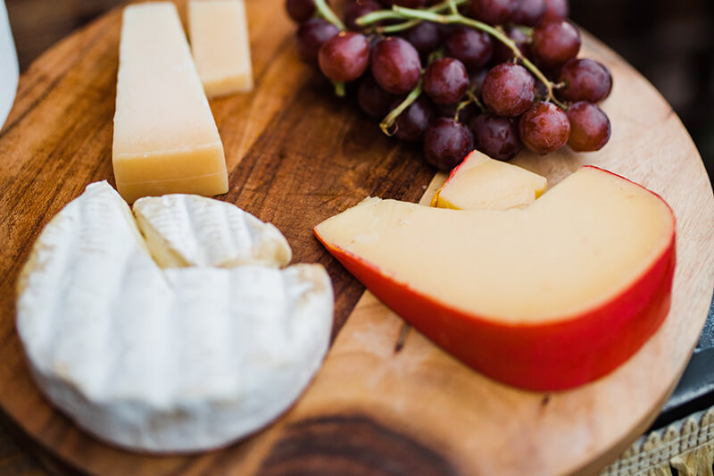 Na drewnianej desce leżą kawałki różnych rodzajów serów razem z ciemnymi winogronami do przegryzania.