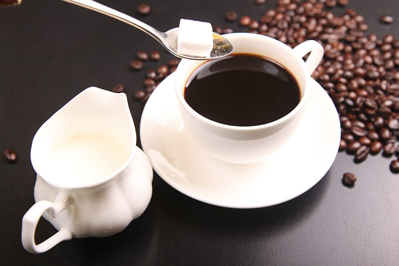 Nad filiżanką kawy czeka łyżeczka z kostką cukru, a obok widzimy dzbanek mleka i ziarna kawy.
