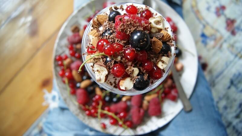Na stoliku leży talerz i pucharek pełen zdrowych przekąsek: kolorowych owoców, orzechów, migdałów, jogurtu naturalnego i kakao.