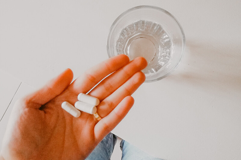 Osoba trzyma 3 białe kapsułki na otwartej dłoni, pod którą widać szklankę wody do ich popicia.