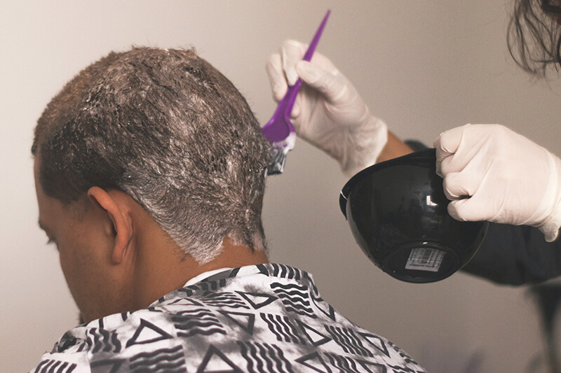 Fryzjerka nakłada ciemnowłosemu mężczyźnie ziołową farbę przy pomocy pędzla i w rękawiczkach zabezpieczających.