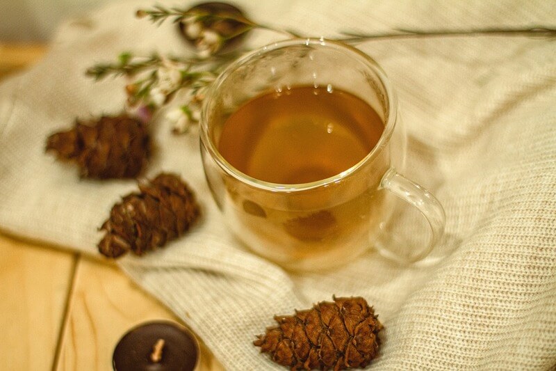 Na naturalnym obrusie stoi filiżanka z sosnową herbatką, w wokół niej leżą szyszki i zioła.