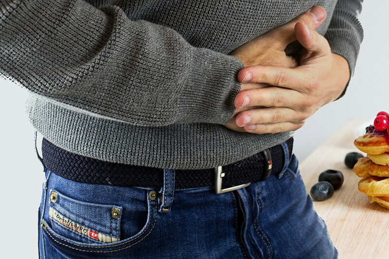 Mężczyzna w szarym swetrze trzyma się za brzuch w skurczonej pozycji sugerującej ból, zgagę lub niestrawność.