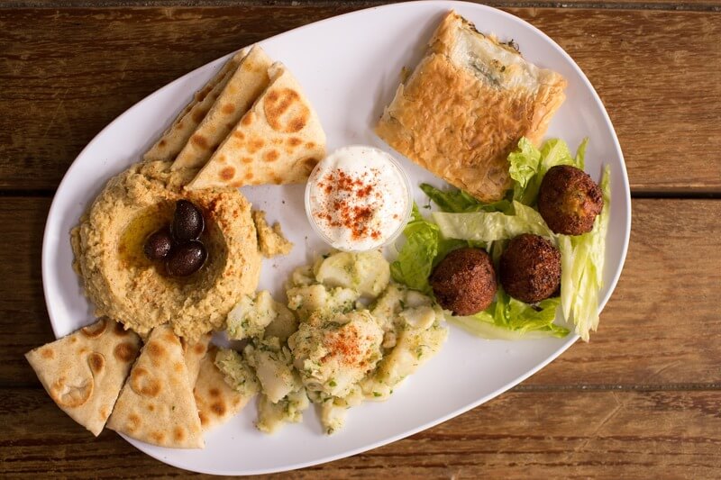 Na szerokim talerzu podano różnorodne dania kuchni arabskiej - chlebek, ciasto, pieczonego kalafiora, klasyczny hummus oraz falafel na sałacie.