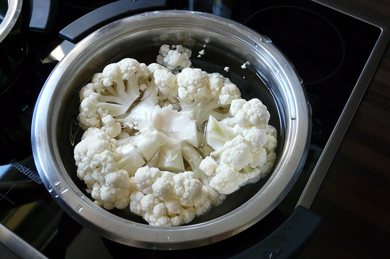 Na płycie indukcyjnej leży garnek z różyczkami kalafiora zalanymi wodą, gotowymi do gotowania.