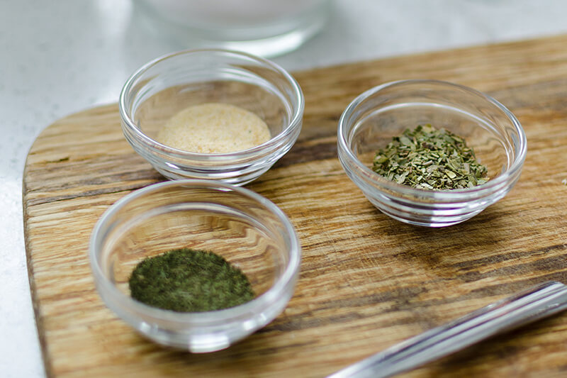 Na drewnianej desce leżą trzy małe salaterki z przyprawami, w tym oregano, czosnkiem i tymiankiem.