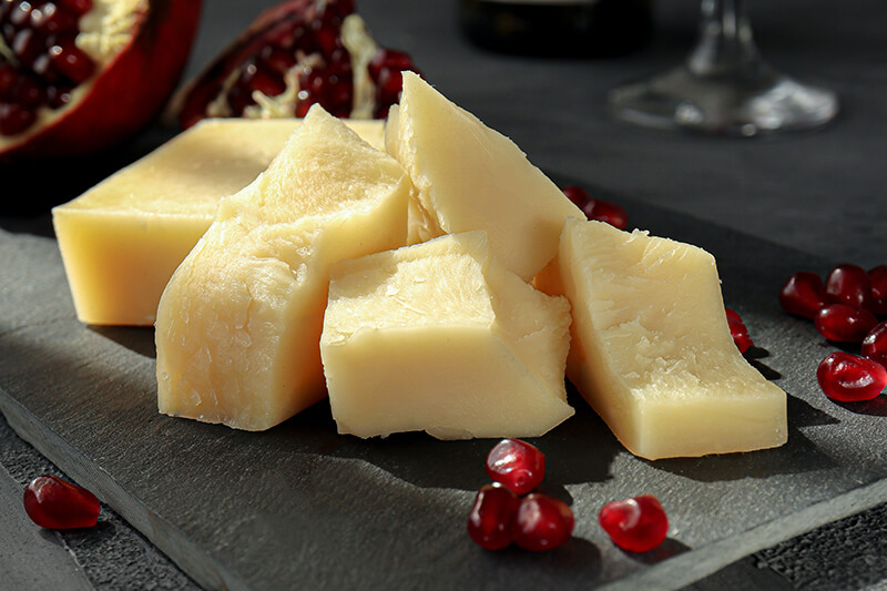 Na czarnej desce leżą kawałki twardego, żółtego sera bez laktozy oraz pestki granatów.