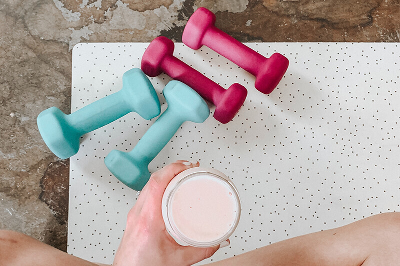 Kobieta siedzi na macie po skończonym treningu, w ręku trzyma szklankę z białkowym jogurtem.