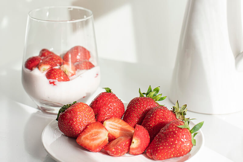 Na talerzu leżą świeże, soczyste truskawki, a obok nich w szklance znajduje się truskawkowy deser z jogurtem.