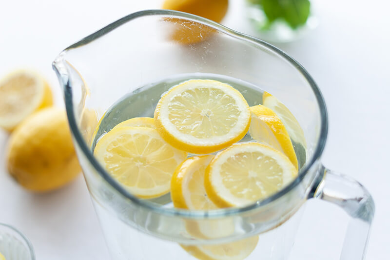 Na stole stoi szklany dzbanek ze świeżą wodą, do której wkrojono plasterki soczystych cytryn.